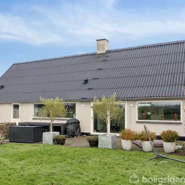 Et enfamiliehus med sort tag står i en have med grøn græsplæne. Træer i krukker dekorerer terrassen foran huset.