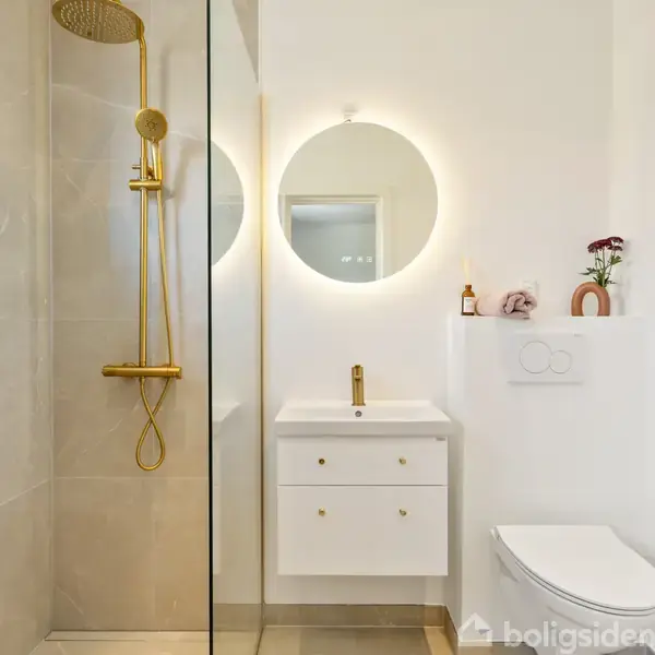 Brusebad med gyldenbruser står ved siden af en glasvæg. En vægmonteret vask med rund spejl og lys over, findes i nærheden af et væghængt toilet. Minimalistisk, moderne badeværelse.