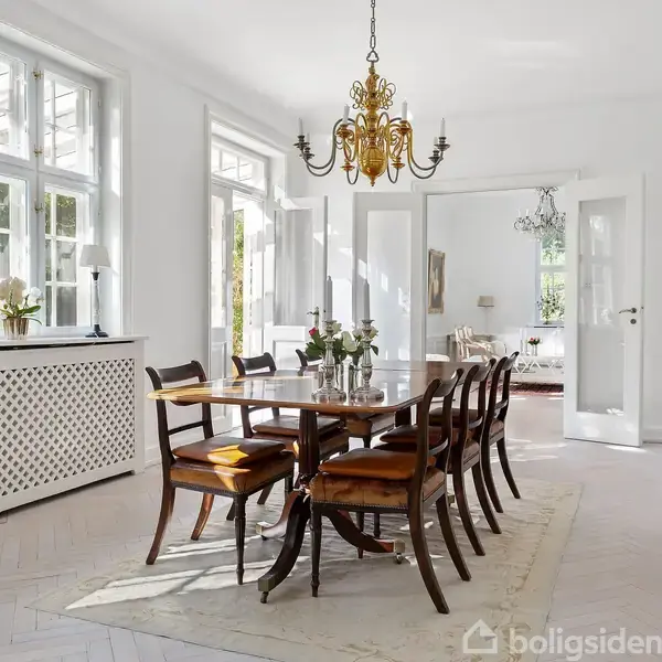 Et elegant spisebord med seks stole står i et lyst og rummeligt spisestue. Over bordet hænger en dekorativ lysekrone. Store vinduer og åbne døre giver adgang til en tilstødende stue.