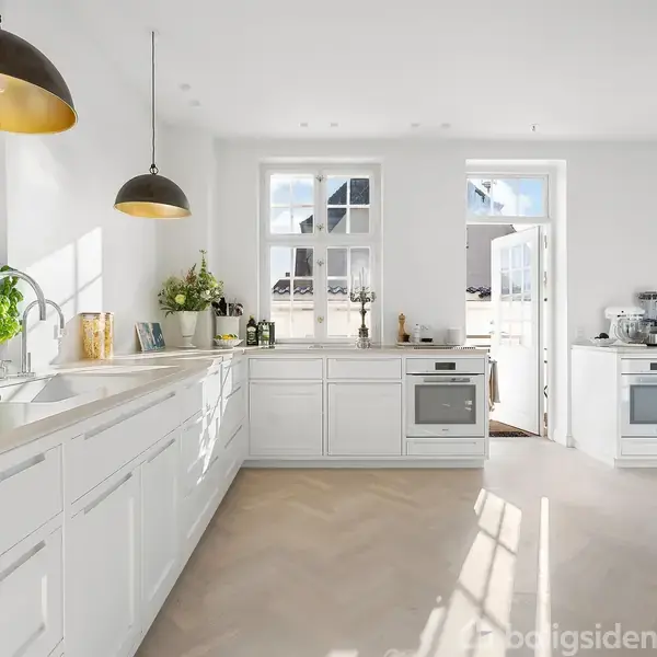 Hvidt køkken med moderne indretning, sollys strømmer ind gennem vinduerne. Køkkenø og borde er dekoreret med planter og køkkenredskaber. Ovn og vask er integreret i skabene.