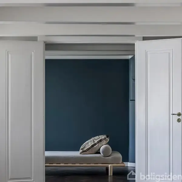 Hvide døre åbner mod et rum med en mørkeblå væg. En stak puder og en rund pude ligger tæt på væggen, hvilket skaber en minimalistisk og stilfuld atmosfære.