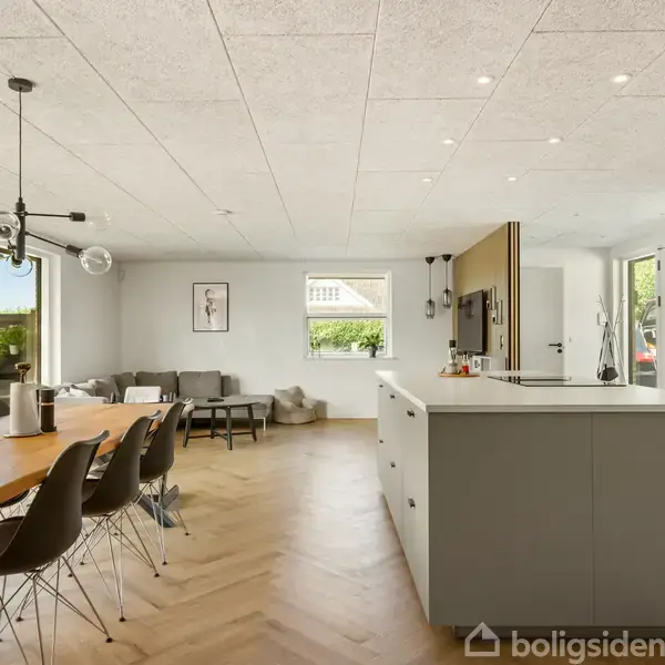 Et moderne køkken-alrum med et stort køkkenbord i forgrunden. Spisebord med sorte stole til venstre, og stuehjørne med sofa og bord i baggrunden. Store vinduer mod haven.