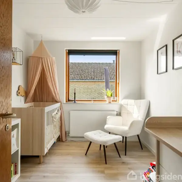 En babyseng med en rosa baldakin står i et lyst rum. En hvid lænestol med skammel står ved et vindue med udsigt til et tag. Væggen har børnedesign.