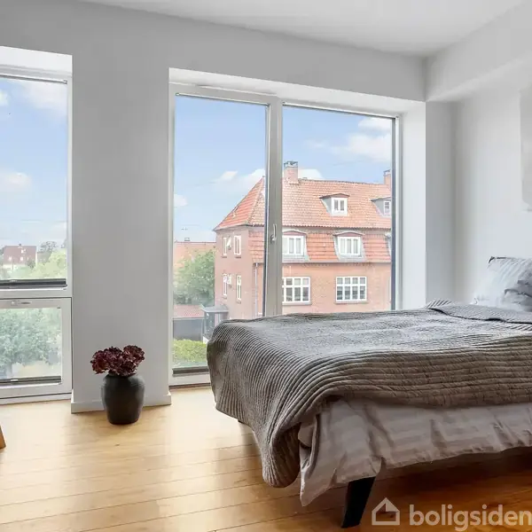 Soveværelse med dobbeltseng dækket af gråt sengetæppe, placeret ved store vinduer. Der er en kommode med lampe og plante. Udsigt til røde murstensbygninger udenfor.