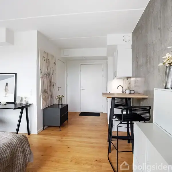 Et moderne værelse med seng til venstre. Skrivebord med stol i midten, og lille køkken med barstole til højre. Lyse træmøbler og minimalistic dekoration.