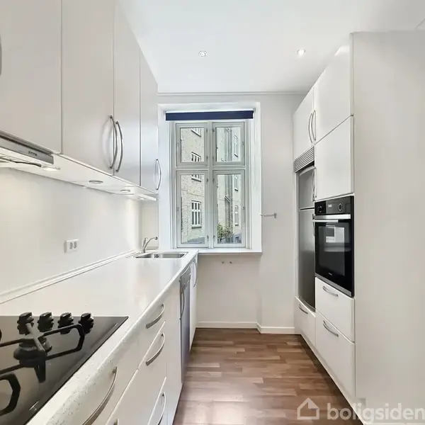Et moderne, smalt køkken med lyse hvide skabe, indbygget ovn og kogeplade. Køkkenet er belyst af naturligt lys fra et vindue for enden. Gulvet er af mørkt træ.