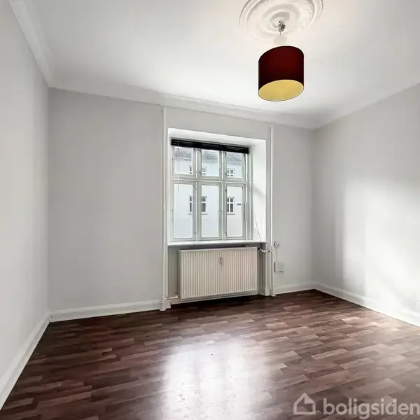 Et tomt, lyst rum med trægulv. En hvid radiator og et vindue vender ud mod gaden. En rød lampeskærm hænger fra loftet, og døren står åben.