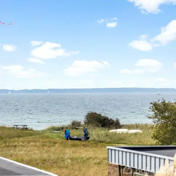To personer bærer en lille båd hen over en græsmark nær en kystlinje. I baggrunden ses havet med sejlbåde og en klar blå himmel. En flagstang med vimpel står i nærheden.