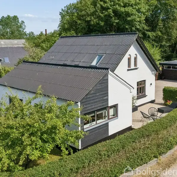 Et hvidt hus med sorte tage står omringet af grønne hække. Foran huset er der et lille terrasseområde med to stole. I baggrunden ses en garage og træer.