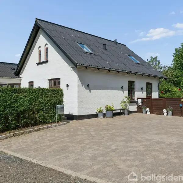 Hvide hus med sort tag, står på en brostensbelagt indkørsel omgivet af hække og træer. Flere potteplanter pryder indgangen, under en klar blå himmel.