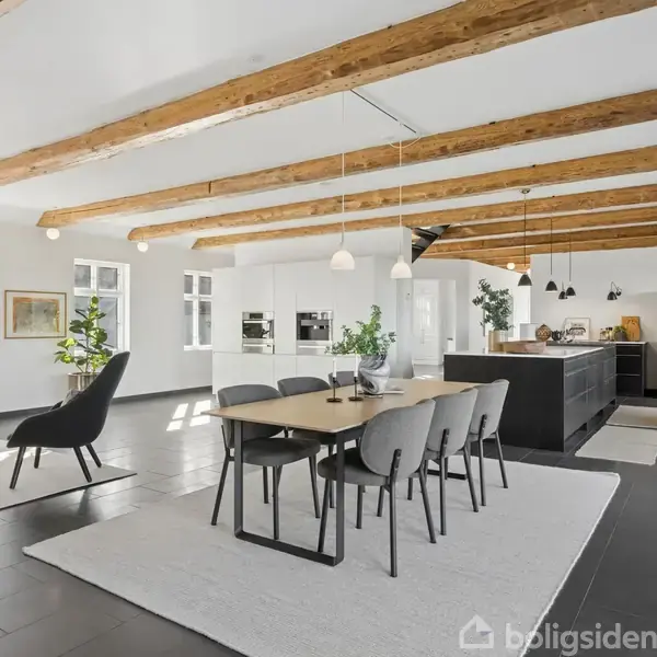 Spisebord med seks stole står på tæppe i moderne, åbent køkken-alrum med loftsbjælker. Køkkenet har ø, hvide skabe og pendellamper. Store vinduer giver naturligt lys og planteliv supplerer indretningen.