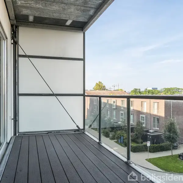 En udendørs balkon med metal- og glasgelænder, med udsigt over et boligområde med træer og parkerede biler i baggrunden, under en klar blå himmel.