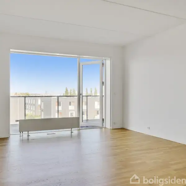Et lyst, tomt værelse med parketgulv har en åben dør ud til en balkon med udsigt over en blå, klar himmel. Væggene er hvide, og rummet er minimalistisk indrettet.