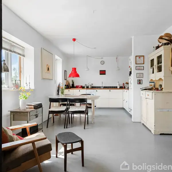 Køkken med hvidt interiør; bord med stole i midten; rød lampe hænger over bordet; vindue med planter til venstre; møbler, billeder og dekorationer i baggrunden.