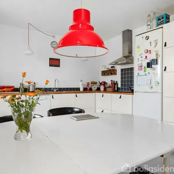Rundt spisebord med en vase blomster står centralt. En rød lampe hænger over bordet. Baggrunden viser et hvidt køkken med skabe, opvask, og emhætte.