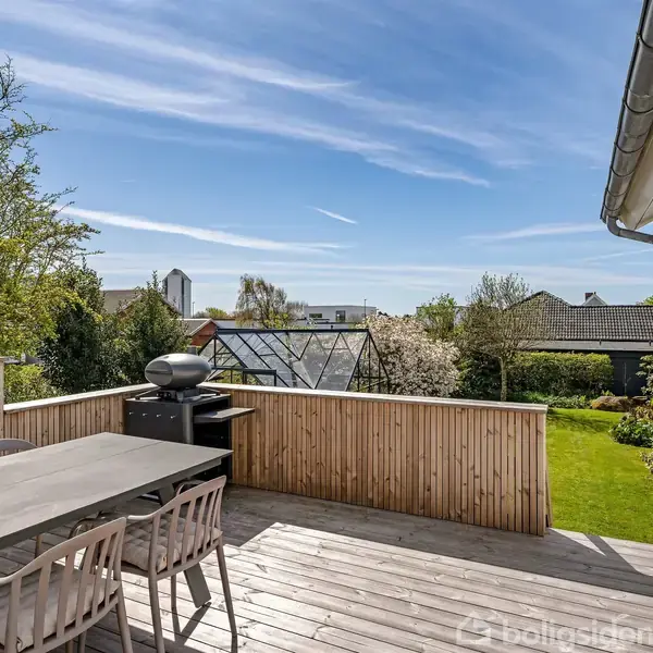 En terrasse med et bord og stole står stille i solen. I baggrunden ses en have med et drivhus og frodige træer under en blå himmel.