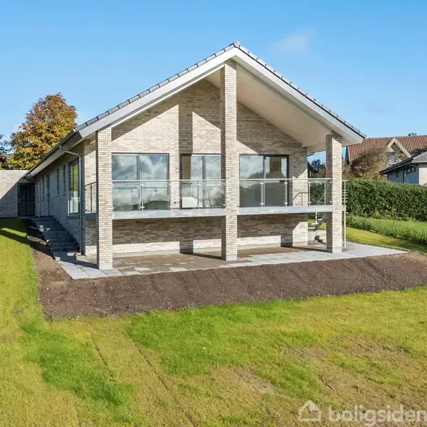 Et moderne hus med store glasvinduer og en overdækket terrasse står i en velplejet have. Hække omkranser plænen, og nabohuse ses i baggrunden under en klar, blå himmel.