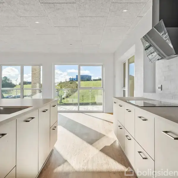 Et moderne køkken med hvide skabe og skuffer står i dagslys. Der er en indbygget vask og emhætte. Store vinduer og en glasdør giver udsigt til en grøn bakke.