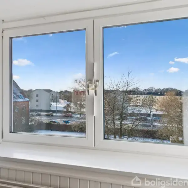 Et vindue viser en udsigt over et boligområde med sneklædte tage og træer, blå himmel med få skyer i baggrunden.