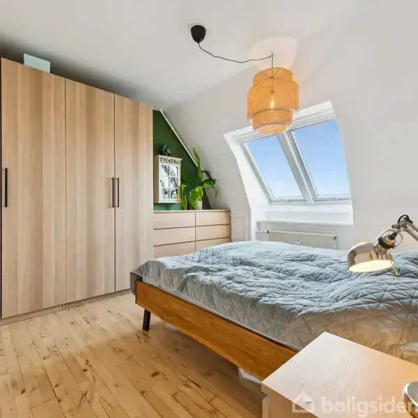 Soveværelse med en opredt seng under et skråvindue, et skab og en kommode. Rummet er lyst med trægulve og en dekorativ lampe. En plante står på kommoden.