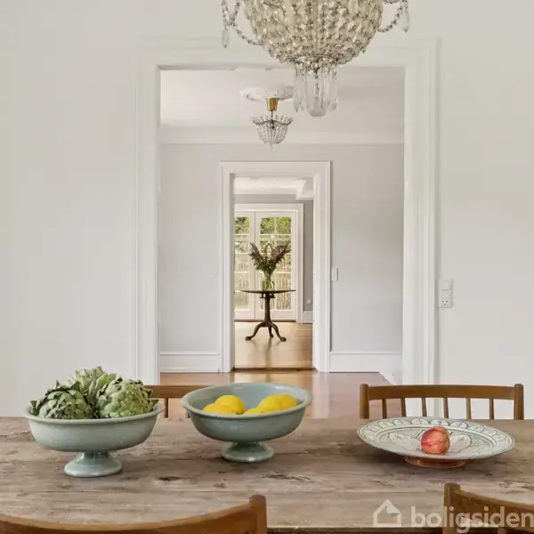 Et rustikt spisebord med skåle af grøntsager og frugter står i et elegant rum. I baggrunden ses en flot lysekrone og en anden stue med en vase og grønne omgivelser.