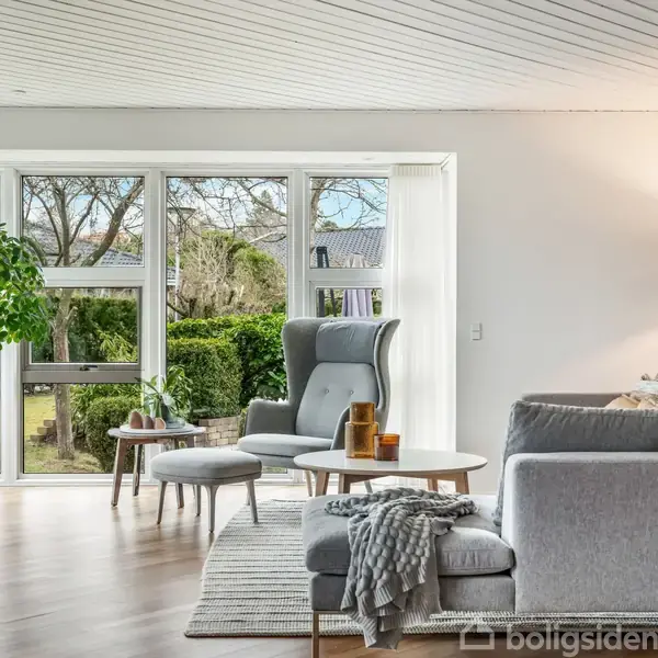 Stue med sofa og lænestol foran store vinduer, som viser en have. Et tæppe og et sofabord med dekorationer er placeret ved siden af sofaen, og en stor plante står i hjørnet.