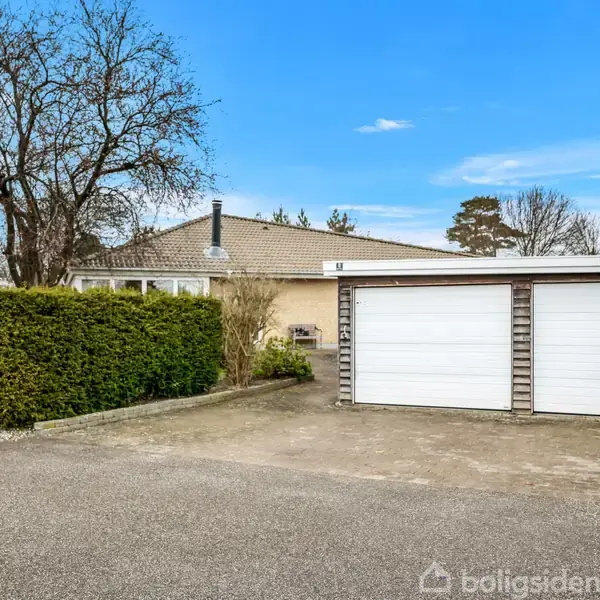 Et parcelhus med fladt tag og en tilhørende garage med to hvide porte, omgivet af grusbelagt indkørsel og grønne hække. Fyldigt træ står i haven. Ingen synlig tekst.