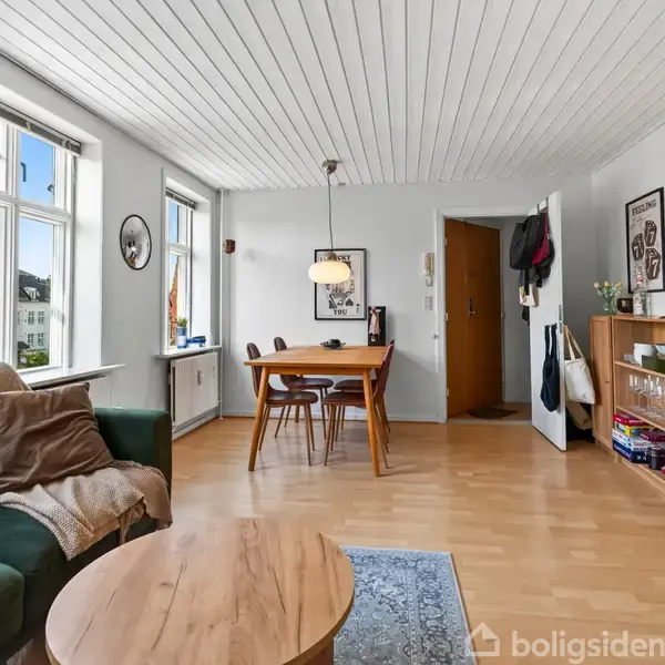 Grønt sofaarrangement med puder, bord og tæppe; spisestue har træbord og stole. Væggene har dekorationer, og der er store, lyse vinduer. En reol og en stor spejl står ved siden af døren.