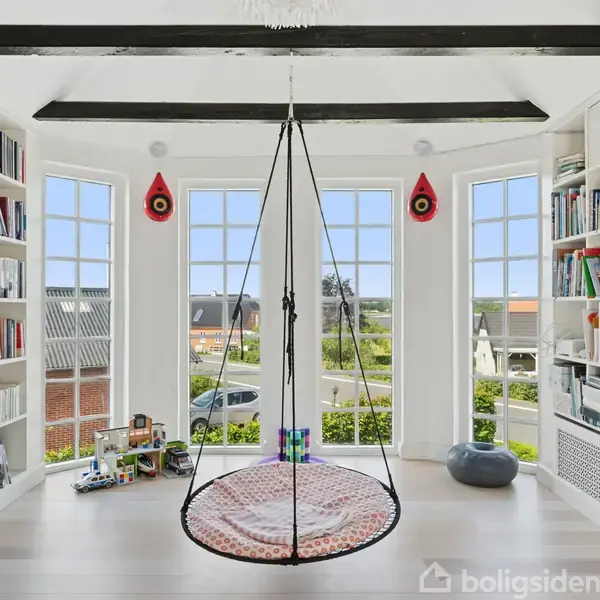 Et rundt, hængende netgynge svæver foran store vinduer med udsigt til et nabolag. På begge sider er der reoler fyldt med bøger og små dekorationer.