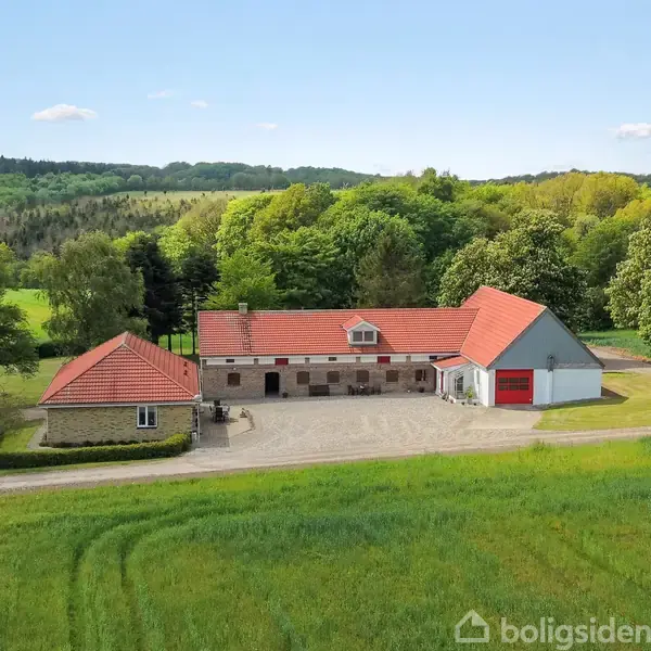 En gård med tre røde tegltage bygninger står stille i en frodig, grøn natur omgivet af skov. Grusindkørsel fører til en velholdt plads foran bygningerne.