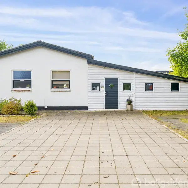 Hvidt hus står roligt omgivet af træer og en flisebelagt indkørsel. To vinduer og en grå dør med et lille vindue præger facaden.