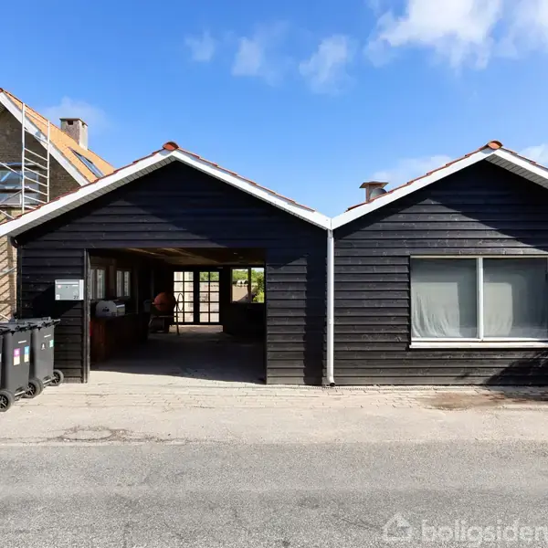Hus med sort træfacade og to garager i midten, omgivet af hække og skraldespande i forgrunden; et større hus under renovering ses bagved.