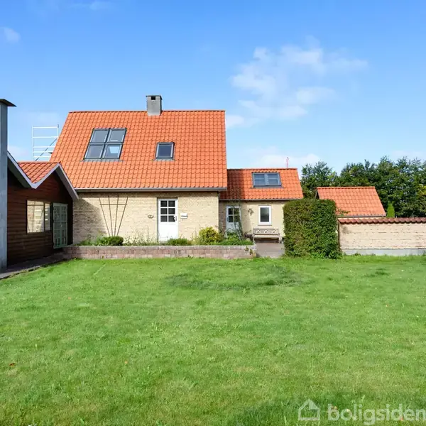 Et hus med rødt tegltag og flere vinduer står midt i en grøn have, flankeret af en mindre, træbeklædt tilbygning. En blå himmel er i baggrunden.