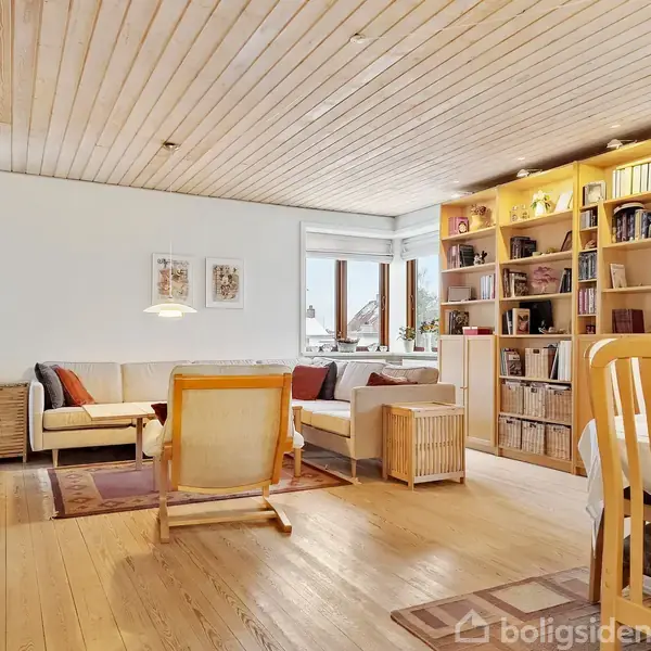 En stue med sofa og lænestol omkring et tæppe, omgivet af hylder fyldt med bøger. Et spisebord med stole står til højre, og vinduer lader naturligt lys ind.