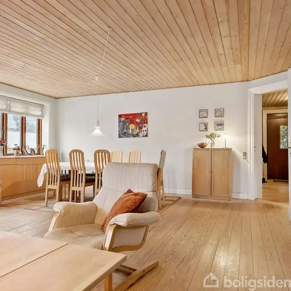 En stue med trægulv og træloft har en sofagruppe og en lænestol. En bogreol fyldt med bøger står ved siden af et spisebord med stole. Sollys strømmer ind fra vinduerne.