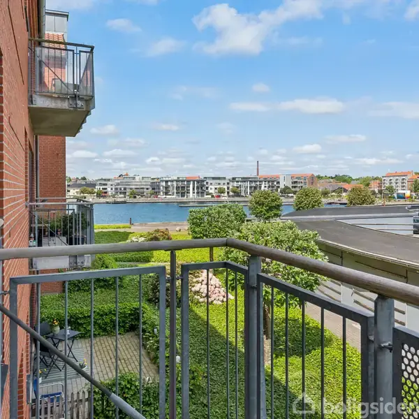 En balkon med metalrækværk i en murstensbygning giver udsigt over en vej, grønne buske, og en sø eller flod med moderne bygninger i baggrunden under en blå himmel med skyer.