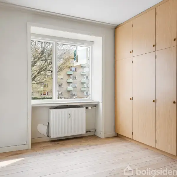 Et lyst værelse med trægulv. Indbygget klædeskab fylder højre væg. Et vindue med hvid ramme viser udsigt til lejlighedsbygninger og træer. Radiator under vinduet.