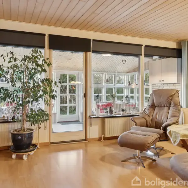 En stue med lænestol og fodskammel ved siden af et stort stuevindue med udsigt til veranda. En grøn plante står ved glasset. Møblerne er brune, og gardiner hænger på siden.