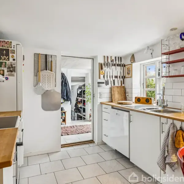 Et lyst køkken med hvide skabe og trækøkkenbord, organiseret med køkkenredskaber på væggen og hylder. En dør åbner ud mod en anden lys, indrettet stue. Ingen tekst ses.