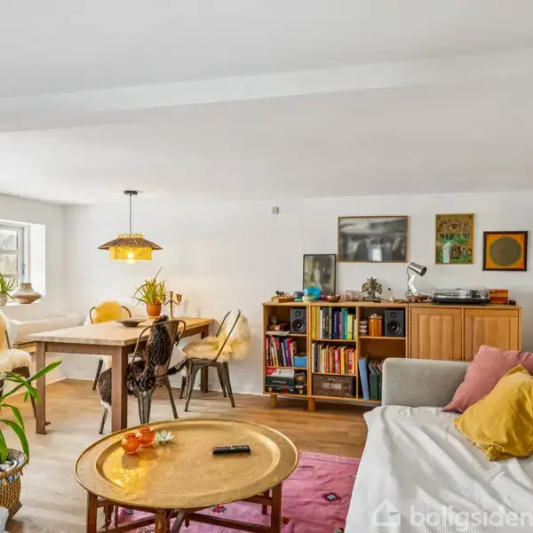 Stue med sofa og rund stuebord med frugt og fjernbetjening, ved siden af et TV på væggen. Baggrund: spisebord, stole, reol med bøger og dekorationer, lyse farver.