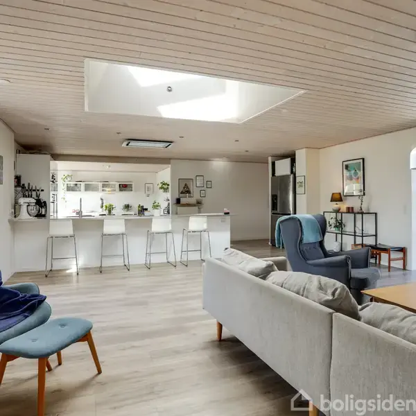 Stue med sofa og stol står ved et sofabord. Baggrunden viser et lyst køkken med barstole. Store vinduer slipper dagslys ind, skaber en åben og indbydende atmosfære.