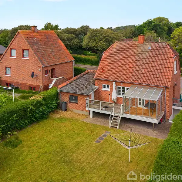 Et rødt murstenshus med en stor have og terrasse. Huset har en glastilbygning. Det er omgivet af grønne hække og flere andre murstenshuse i en villakvarter.