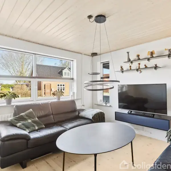 Lædersofa og rundt sofabord står foran et stort vindue i en stue. Et fladskærms-tv er monteret på væggen, med hylder ovenover, og der er en moderne lampe i loftet.