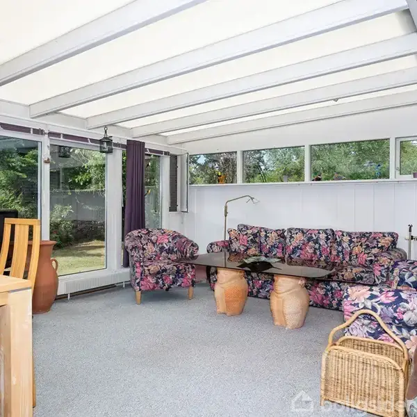 Sofa med blomstrede puder står i en lys udestue med glasvægge og loft. Der er et sofabord med keramiske krukker som ben og en flettet kurv ved siden af sofaen. En træstol står ved et spisebord.