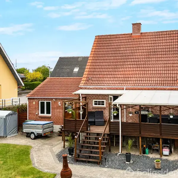 Rødt murstenshus med tagterrasse og trappe op til veranda. Campingvogn står til venstre, og der er en have i forgrunden. Nabohus og veje ses i baggrunden.