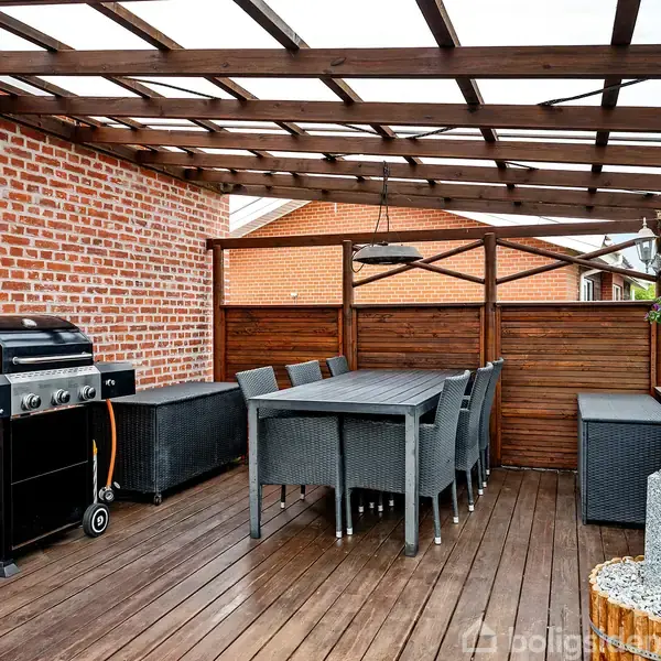 Grill står på træterrasse under pergola. Havebord med stole ved siden af, omgivet af murstensvæg og træhegn. Terrasse dekoreret med planter og stengruppe.