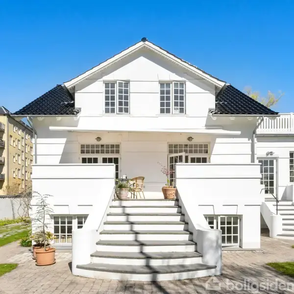Hvid villa med sort tag står midt i en have. Trappe fører op til indgangen, omgivet af potteplanter. Solrig dag med blå himmel i et byområde.