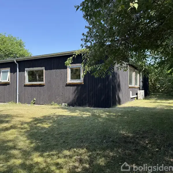 Et sortmalet hus står stille i en solrig have, omgivet af grønne træer og en velplejet græsplæne under en klar blå himmel.
