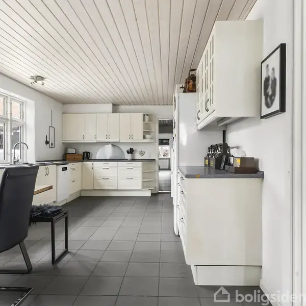 Køkken med hvide skabe og sort bordplade. Rummet har et spisebord med sorte stole. Store vinduer giver lys, og en dør åbner mod en stue med en grøn plante.