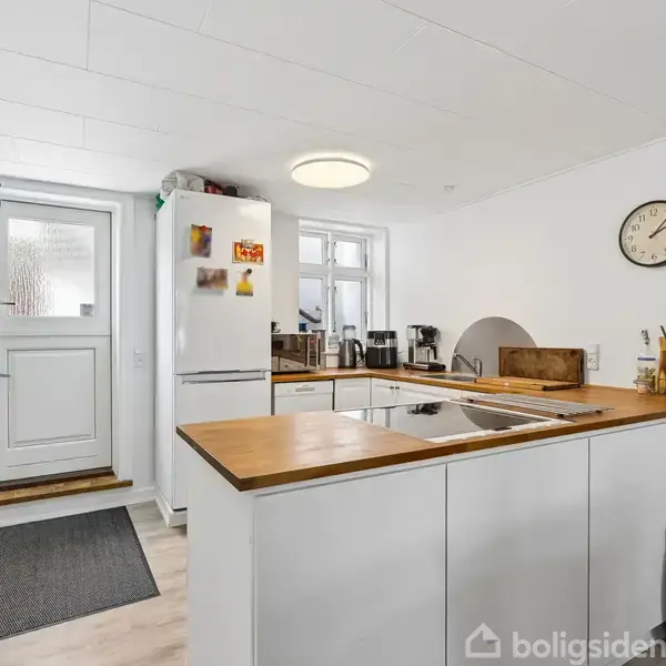 Køkken med hvide skabe og træbordplade; køleskab ved dør med tøjstativ; væghængt ur viser klokken 3:50; skohylde nær sort måtte på gulv.